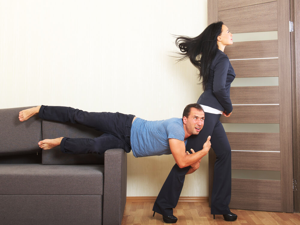 A man hanging off a couch, clinging desperately to a woman's leg as she leaves for work.