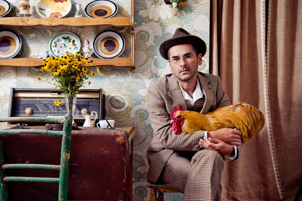 A man wearing a vintage suit and hat, surrounded by vintage furniture and holding a chicken.