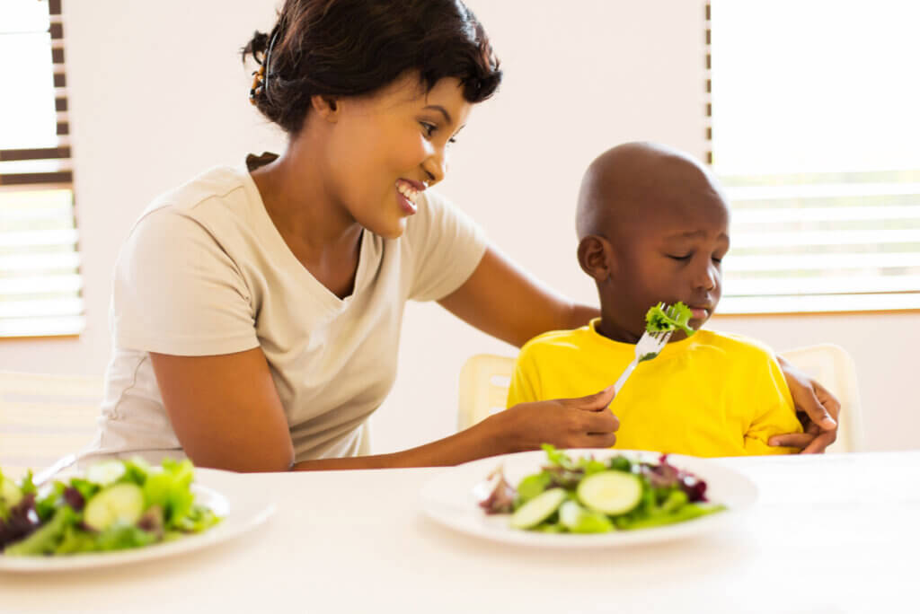 A black child turns away from his mother as she offers him a fork full of vegetables.