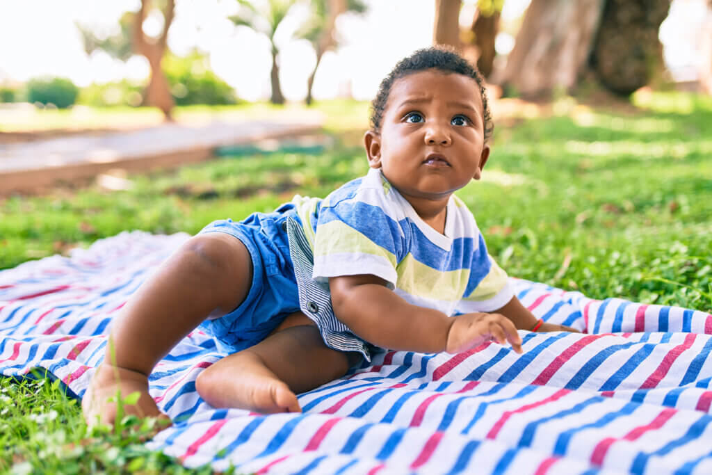A black baby sitting on a blanket in the grass.