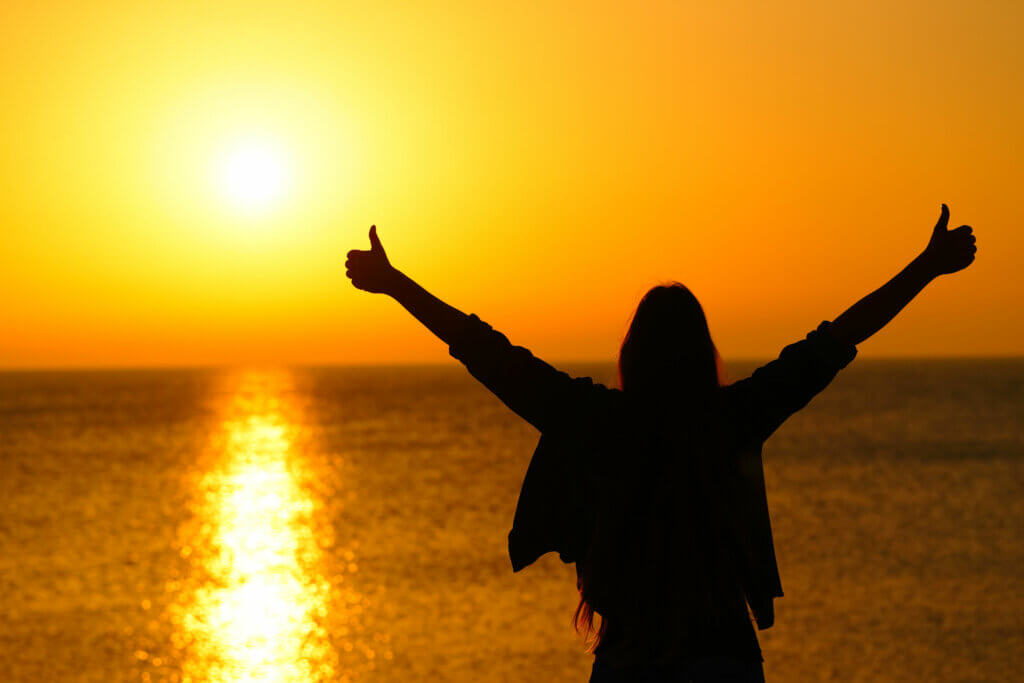 A woman looking out at the ocean at sunset with her hands in the air, making two thumbs up.