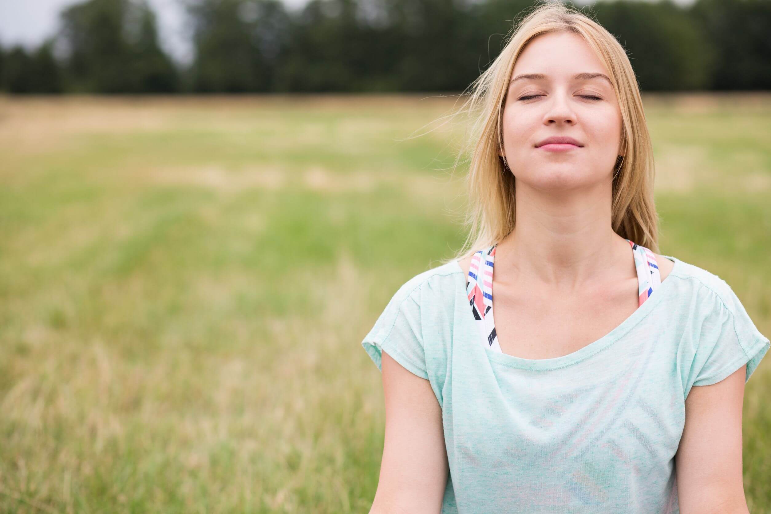 A young woman smiling as she meditates in a field.