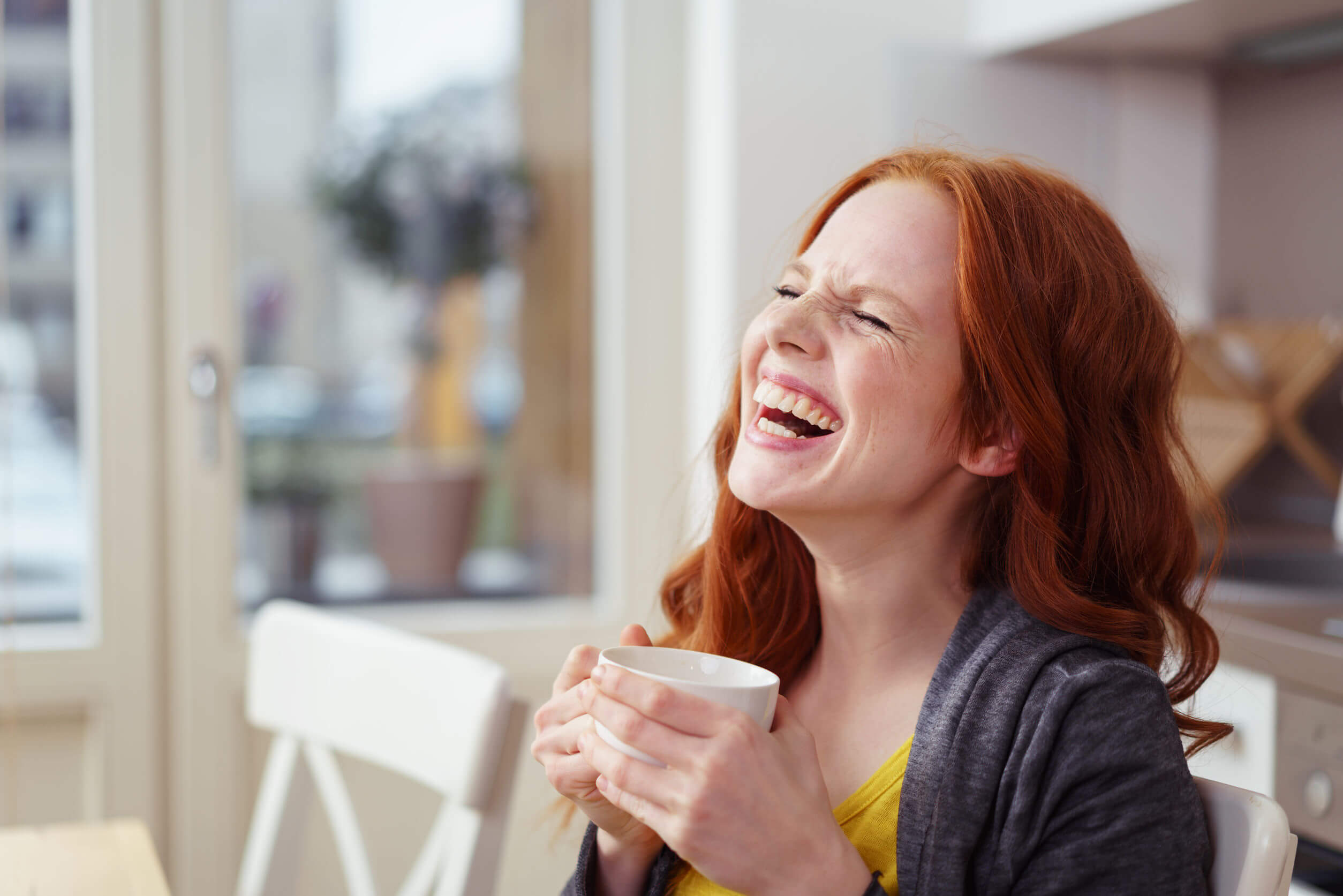 Á red-headed woman sitting at a table, holding a cup of tea, and laughing heartily.