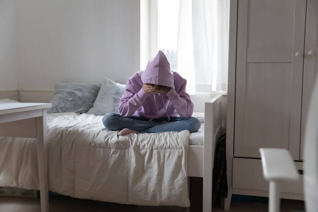 Une personne assise sur un lit cachée dans sa capuche.